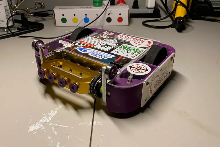 A purple and gold beetleweight combat robot on a workbench next to soldering equipment.