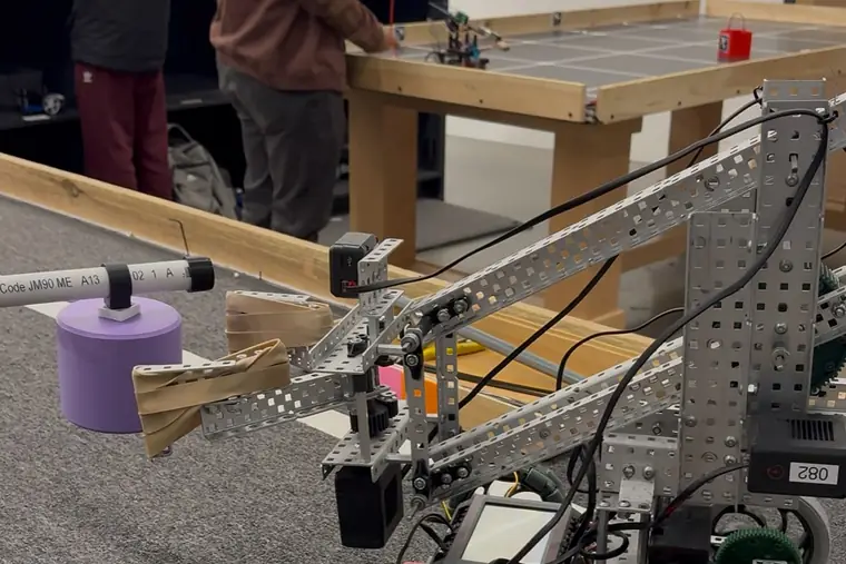 A VEX robot extending its arm to grip a purple cylindrical fruit from a wooden branch.