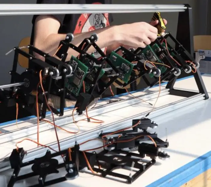 The Shive wave machine part-built on a workbench: a row of pendulum arms
              carrying circuit boards mounted along a torsion rail, with further arm
              assemblies laid out in the foreground.